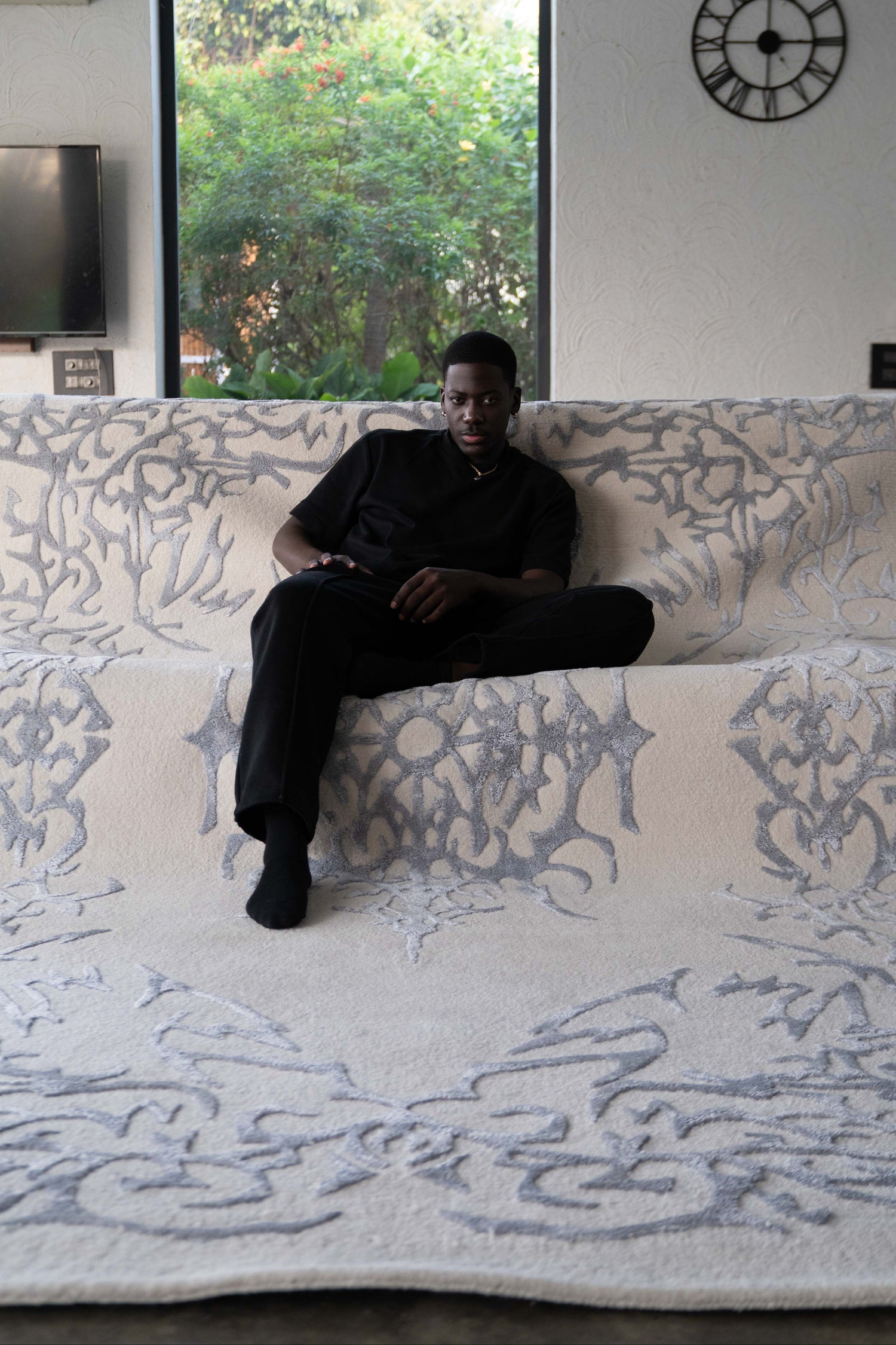Man sitting on a large patterned rug in a room with a clock on the wall.